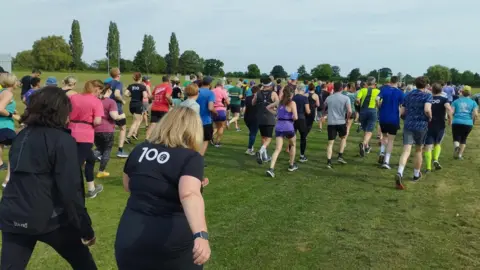 A group of runners of varied ages and attire participate in a community running event on a grassy path in a park. Some wear running vests and athletic gear, while others are dressed casually.