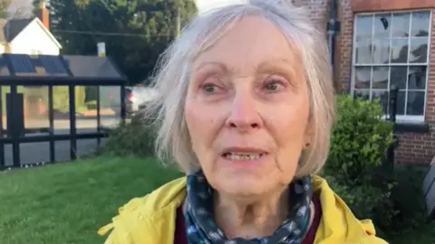 BBC Janette Ward, wearing a yellow jacket, stands in front of her local bus stop which she uses to catch services to see her daughter and go to appointments