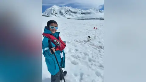 Yuam (left) smiling at the camera has a turquoise jacket on with red life jacket around his neck and sunglasses is holding onto a walking pole and pointing at a penguin.