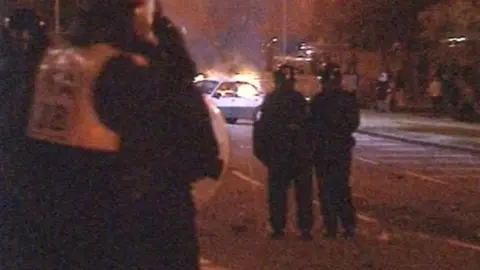 A grainy photo from 1995 showing police officers wearing armour and stood together looking at a car on fire.