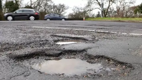 Two large potholes on a road, which has cars being driven on it in the background.