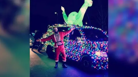 Scott Dickens A  man dressed as Santa smiling and posing in front of a van decorated all over with rainbow fairy lights. On top of the car is a lit-up inflatable dinosaur.