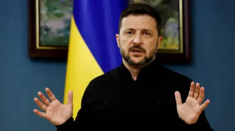 Reuters O presidente da Ucrânia gesticula com as mãos em um top escuro com uma bandeira azul e amarela ucraniana atrás dele
