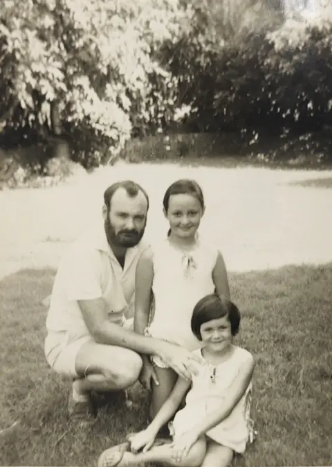 10 -year -old Michelle Dujerti stands in a black and white image of 1972 with her father to the right and her younger sister in the foreground. It appears to be in a sunny garden and wear white or bright sleeves.