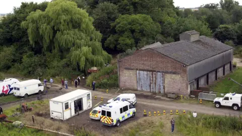 Two police vans, police car and a police mobile stand in front of a sprawling modern barn, with police officers walking to a gate, and members of the media with TV cameras close by. Towering trees surround the site.