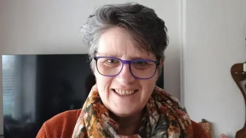 A woman with a short grey heair and purple glasses is standing in front of a large black TV screen and smiling at the camera