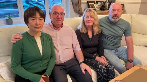 Bo McGenity, Liam's stepmother wears a green cardigan. Peter McGenity, 65, Liam's father wears a striped t-shirt and glasses. Helen Price, 65, Liam's mother, wears a short cardigan and Alan Price, 67, Liam's stepfather, wears a green T-shirt. All four are sitting on a cream colored sofa.