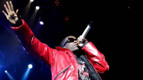Tinchy Stryder performs on stage at night. He is wearing a red coat, a black and red T-shirt, and big sunglasses. He is holding a microphone to his mouth and has his other arm extended. Stage lighting can be seen behind him.