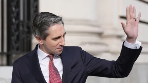 PA Media Simon Harris wearing a dark suit, with a white shirt and a red tie, waving. He is standing in front of a government building. 