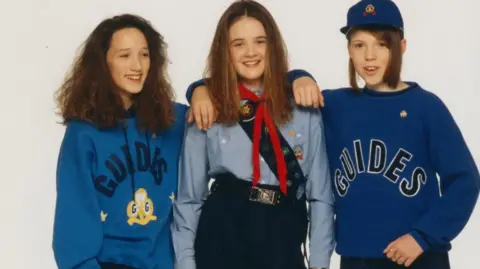 PA Media Three girls standing next to one another and smiling with their arms around each other. The girl on the left is wearing a blue hoodie with the word 'Guides' on it in navy text above a yellow and white Girlguiding logo. The girl in the middle is wearing a light blue shirt, red neckerchief, and badge sash. The girl on the right is wearing a royal blue sweatshirt with 'Guides' on it in large letters and a cap.