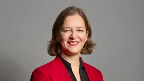 UK Parliament Parliamentary photo of Fleur Anderson wearing a red jacket and black shirt stood in front of a grey background