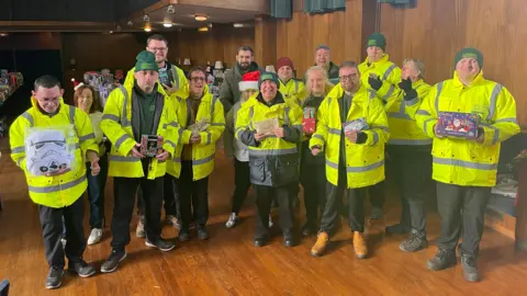 Sefton Council A large group of people wearing high viz yellow coats holding a selection of toys to camera and smiling ins ide Bootle Town Hall