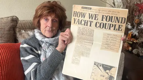 BBC Pat Brewin sat on a sofa holding a newspaper cutting. Pat is 79 years old and has short, dark hair. She is wearing a grey jumper with white horizontal stripes, and a light grey scarf with butterflies on. 