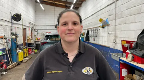 Amelia Riley/BBC News A woman with brown hair tied into a bun stands inside a garage, looking into the camera. She is wearing the same black T-shirt as the man, with the company branding in yellow.