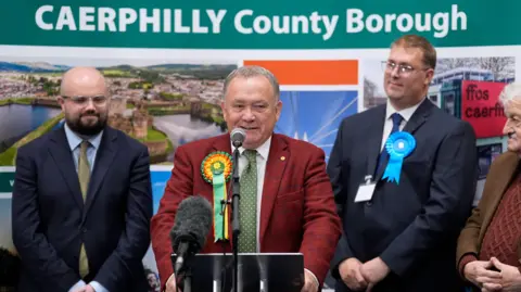 PA Media Plaid Cymru's Lindsay Whittle makes a speech after being declared winner for the Caerphilly Senedd by-election, at Caerphilly Leisure Centre. Reform's Llŷr Powell is to his right, with Conservative Gareth Potter and the Green Party's Gareth Hughes to his left.