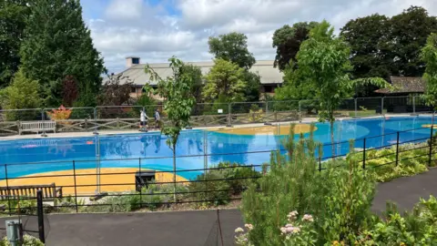 A public paddling pool with blue and yellow flooring. It is surrounded by a footpath and trees