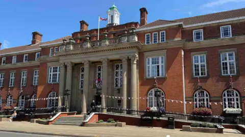 Image showing the exterior of Nuneaton and Bedworth Borough Council's headquarters. The building is red brick with lines curved windows on the ground floor and rectangular windows on the two floors above. All feature white frames. There are steps leading to the main entrance which features six distinctive sandstone pillars which extend from the ground to above the second floor. A single Union Flag flies from a flagpole above the entrance.