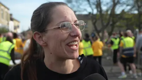 A woman smiling during an outdoor event. She is wearing glasses, has a nose piercing and has her hair is tied back. She’s dressed in a dark top and is speaking into a microphone. In the background, people in yellow T‑shirts and hi‑vis vests are gathered.