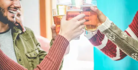 Group of friends toasting each other with beers in a pub. Four arms can be seen in green and red tops, with the focus on close-up glasses.