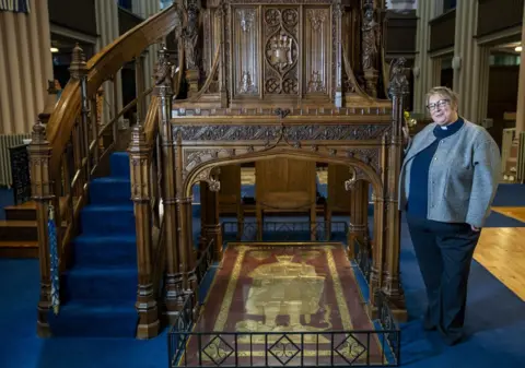 PA Media Rev Dr Mary Ann Rennie, wearing navy trousers and top with a clerical collar and grey cardigan, stands in the Abbey Church next to the tomb of King Robert the Bruce, with a brass image of the king inlaid into red stone with a carved wooden surround. 