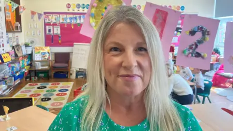Julie is smiling and looking into the camera. She has blond hair and is wearing a green floral dress. She's standing in a classroom. There are primary school children sitting behind her.