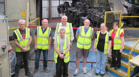 Leafield Environmental Limited A group of staff in high-vis in a factory all looking up at the camera. Complicated machinery and pipework behind them.