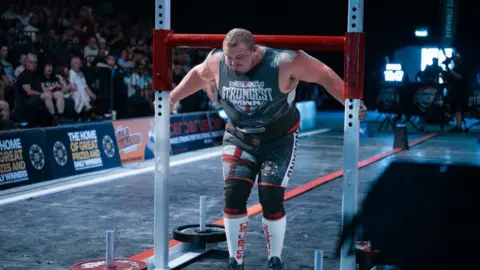 Ben Musgrove Dean Mason - a man with broad shoulders and muscles - is wearing a grey shirt that says England's Strongest Man, shorts with the St George's flag on them and knee-high white socks. His face shows he is straining while trying to push metal bars along the floor. A crowd can be seen to the left of him.