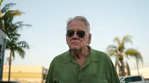 BBC/Passion Pictures Ltd/James Incledon Gerry Wills who has white hear and is wearing sunglasses and green shirt. Behind him are palm trees and parked cars. 