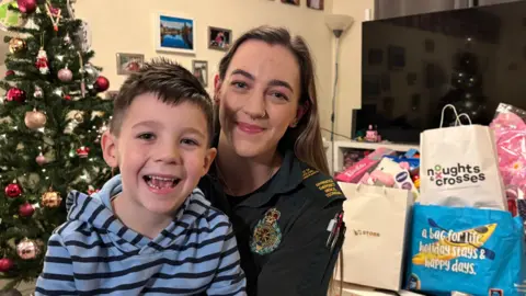 BEN SCHOFIELD/BBC Polly Hurst, with her son Caden sitting on her knee. Both are smiling and looking down the camera. Polly is wearing her East of England Ambulance Service uniform, while Caden has a blue and navy blue striped hooded top on. Polly has long, straight brown hair. Behind them on the left of frame is a Christmas tree, while on the other side is a pile of bags, containing donated toys. 