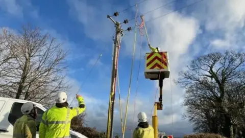 BBC Three workers successful  precocious   vis covering  basal   beside a achromatic  van parked adjacent  to a telegraph poll, which has a cherry picker beside it. Another idiosyncratic    is reaching up   towards the wires.