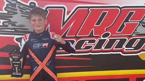 Ste Collinson Jack Collinson, in a navy blue and orange one-piece racing suit, smiling and pointing to a trophy. A red, black, white and yellow banner behind him reads 'MRG Racing'.