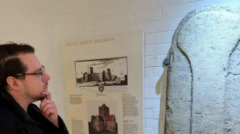 A man with glasses looks at a piece of art. Next to it is a board that says 'anglo saxon religion' 