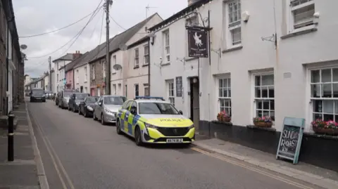 West Street. It is a quiet narrow lane lined with houses and on one side a row of cars are parked, with a police car parked at the front of it. There is a pub called The Thirsty Scholar that has window boxes of flowers and a chalkboard advertising a Sunday roast.