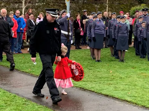 Young girl dressed in a pink dress and red coat carries a wreath of red poppies at a Remembrance Service. She is holding the hand of a man in a black uniform. Cadets and others are gathered behind them.