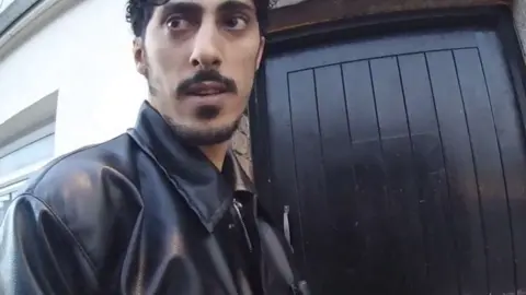 Jersey Police Abdelkarim Arfaoui stands outside a black door in a street. He has short dark curly hair, a beard, and is wearing a black leather jacket.
