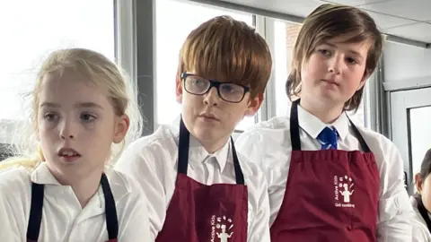 BBC Four primary school children wearing read aprons over their white shirts stand next to each other looking off to their right where their teacher -who's out of shot- talks them through a recipe. They're standing in a bright white kitchen classroom with windows behind them. 