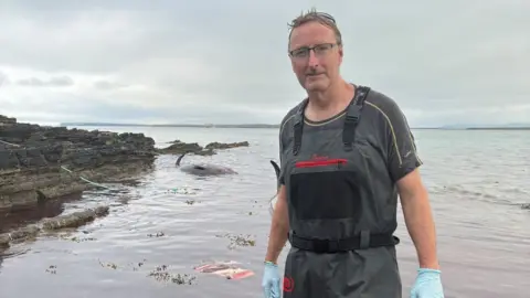 A man stands in front of the sea, looks at the camera. He wears a water resistance and plastic glove. Behind the whale's body is partially covered by water.