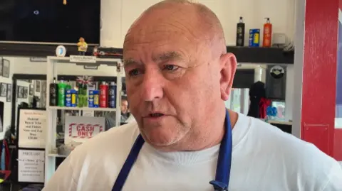 The owner of Steve the Barber talks about the car crash at a nursery. Steve who is bald stands in his barbers wearing a white t-shirt and blue apron straps.
