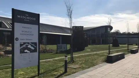 A sign for the World of Wedgewood can be seen in the foreground with the attraction's entrance behind. There is grass and a path leading to the entrance with some benches nearby.