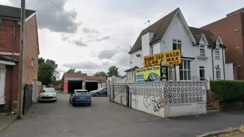 A white building is near the centre of the photo with a yellow sign, with words including hand car wash in black on it, close to the building. Other buildings are on the far left and right of the image.