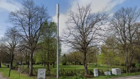 Fordbridge Park in Ashford. The park has a large green field and a number of trees without any leaves. The park is pictured against a blue sky with clouds. A silver telephone mast and other street furniture are in the foreground.