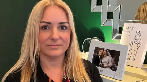 A picture of Caroline Booth, who has long blonde hair, standing in front of a mirror, with a wedding picture of her and her late husband Steve, on the mantlepiece behind her.