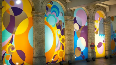 Steven White A multi-coloured series of panels run the length of a tube platform, with geometric shapes and motifs. Some represent plants and others represent circles. 