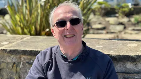 Ken Georgeson has short grey receding hair. He is smiling and wearing sunglasses, sitting in front of a stone wall, with the long green leaves of aa bush behind it. It is a sunny day.