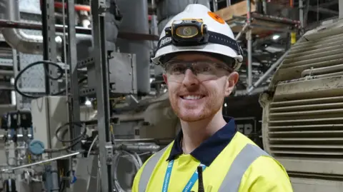 Connor Silom, standing inside the power plant. He is looking directly at the camera and smiling. He is wearing a fluorescent jacket with a blue RWE lanyard round his neck and a white helmet on his head.
