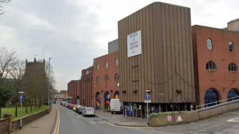 Google The Prince of Wales theatre in Cannock, a modern building with glass doors on the ground flood and other town buildings with murals behind it.