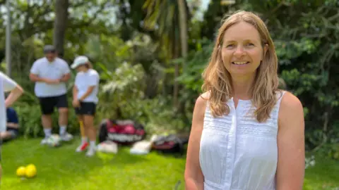 A woman, with shoulder length hair in a white top. She is looking at the camera smiling. She is is stood on the right. In the left in the background are other people and bags with sports balls in them. The grass is vibrant green and there are trees and bushes.