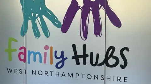 A sign reads 'Family Hubs West Northamptonshire'. The word 'family' is printed out with each letter of the word a different, vibrant, colour. Above the words are two doodles of hand prints.
