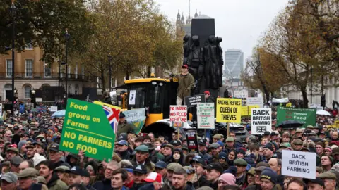 Reuters Farmers take part in a demonstration protesting against the Labour government's new agricultural policy. Some are holding up signs and a yellow tractor can be seen in the background.