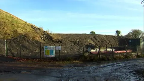 Glan Lash opencast mining site in Llandybie, Carmarthenshire. A mound of land can be seen behind a metal fence with the entrance sign on.